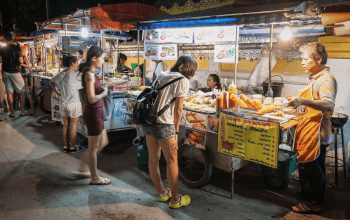 Gambar suasana night market Bangkok yang ramai dengan kios makanan jalanan, lampu warna-warni, dan pengunjung menikmati belanja malam.